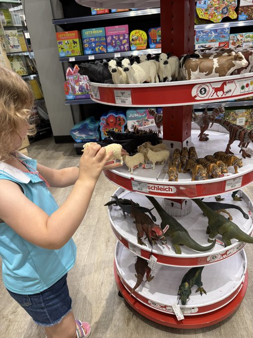 A young girl with blonde hair browsing a circular Schleich toy display at a store, holding a small figurine while looking at the tiered shelves of animal and dinosaur toys.