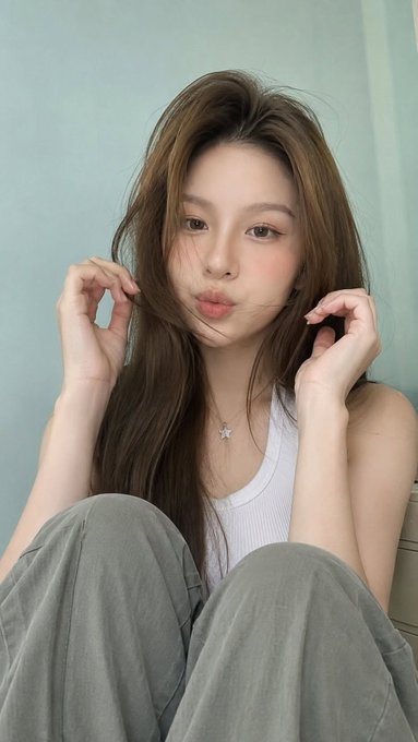A young woman with long brown hair wearing a white tank top and star necklace poses for a selfie, making a cute expression while holding strands of her hair against a light blue wall.