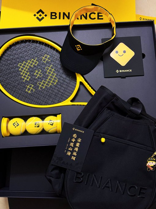 The image features a tennis racket, a black bag with "binance" written on it, and some yellow balls. The items are arranged on top of a table or countertop, suggesting that they might be part of a gift set for someone who enjoys playing tennis. The presence of the tennis racket and the bag indicates that this could be a present for a tennis enthusiast or a sports-related occasion.