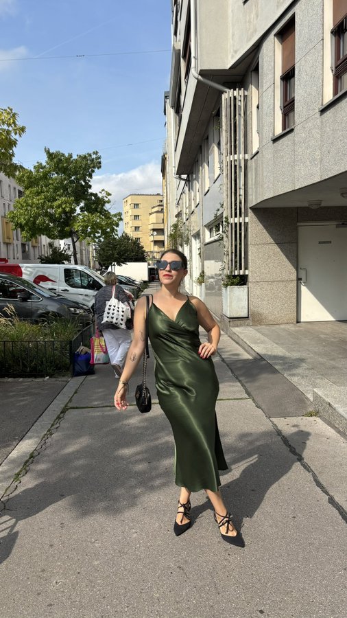 A stylish woman wearing sunglasses and a green silk slip dress standing on a city sidewalk holding a small black handbag, with buildings and parked cars in the background.