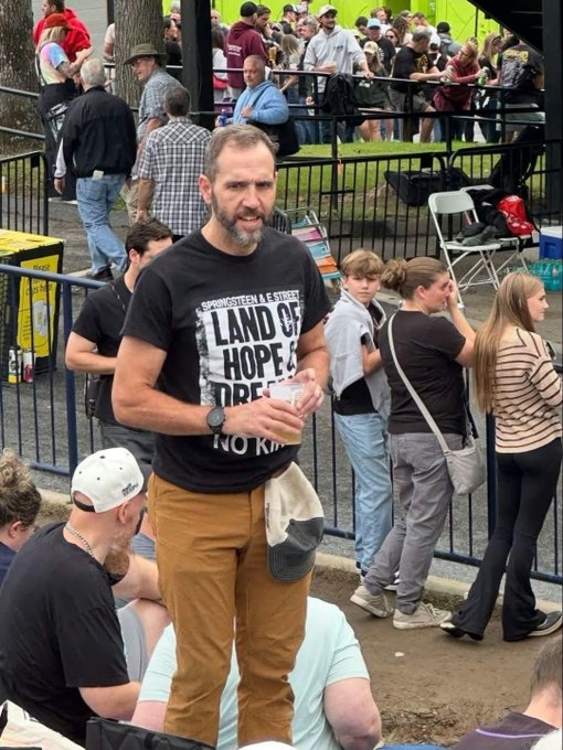 A bearded man wearing a black 'LAND OF HOPE' graphic t-shirt and brown pants stands at an outdoor event with crowds behind a fence, holding a cup and a cap.