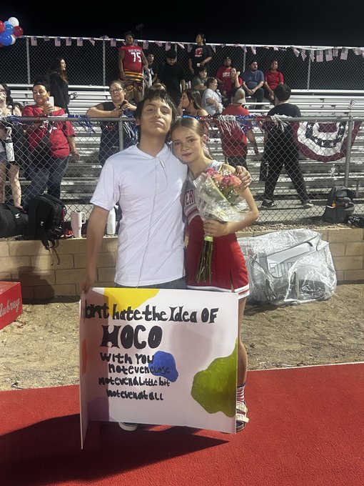 A young man and woman pose together at what appears to be a homecoming event; the man holds a sign reading 'Don't hate the idea of HOCO with your rock, even all the rock, not even all the rock you got all.'