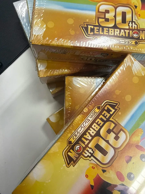 A stack of sealed Pokémon 30th Celebration booster boxes sits on a table, their golden packaging featuring Pikachu and the "30th Celebration" logo with Japanese text. The image showcases the prize won in the lucky draw described in the post.