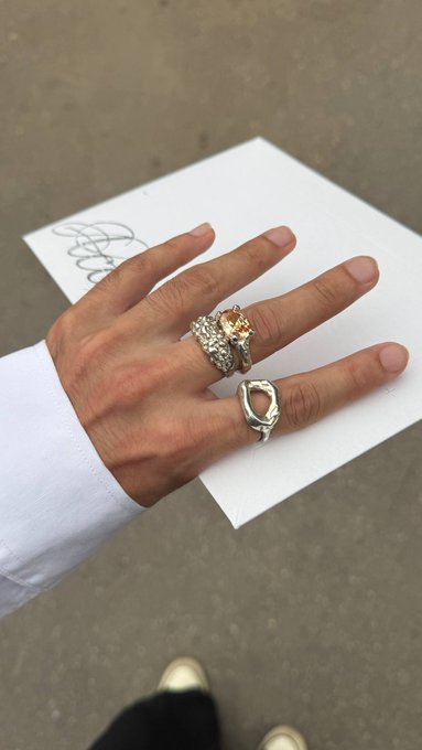 A hand wearing two rings—a diamond-encrusted engagement ring and a sculptural silver band—is held out over a white piece of paper with a signature.