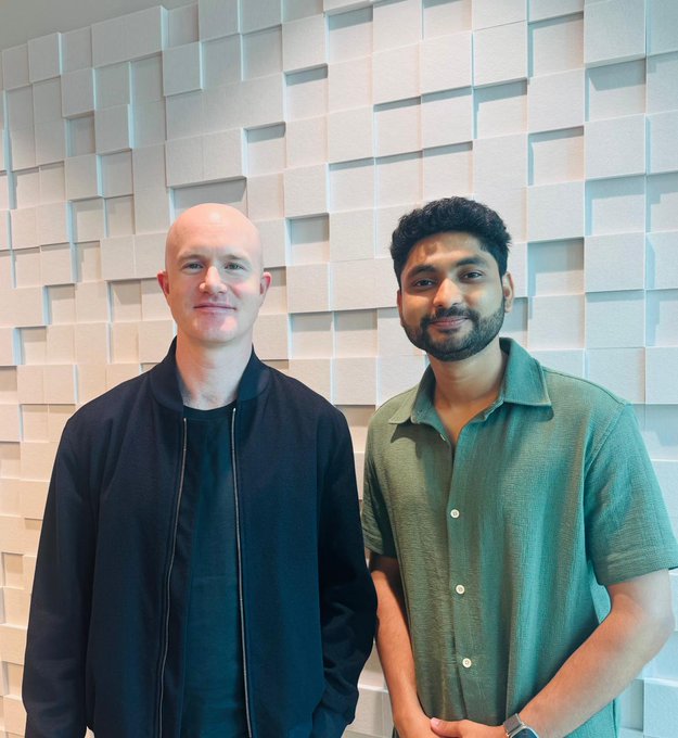 Two men posing for a photo in front of a textured white geometric wall: a bald man in a black bomber jacket and a bearded man with dark hair wearing a green short-sleeve button-up shirt.