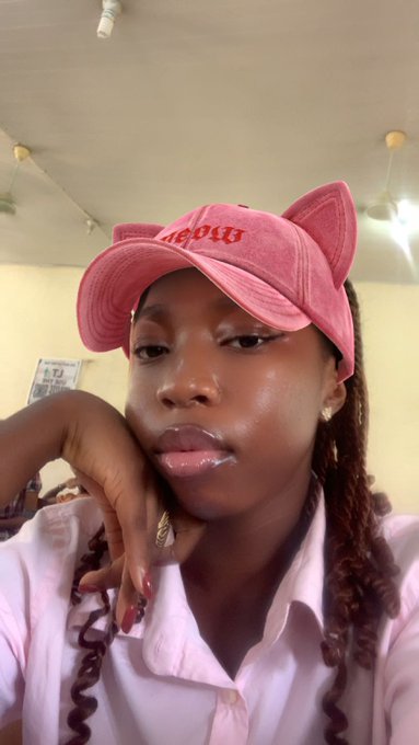 A selfie of a person wearing a pink cat-ear cap with 'Meow' text, a pink button-up shirt, and gold earrings, resting their hand near their face.