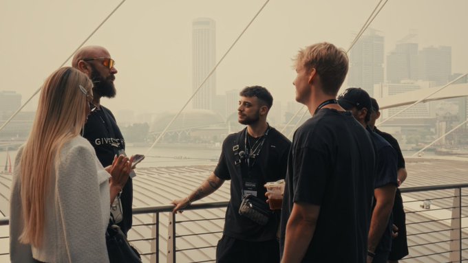 A diverse group of friends chat and check their phones on a bridge overlooking a hazy city skyline, embodying a casual "Good Morning" vibe. One person wears a shirt with partial "GIVEB" text visible, adding a charitable undertone to the gathering.