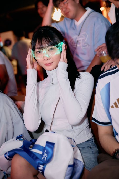 A young woman in a white zip-up jacket poses with glowing green shutter shades while sitting in a crowd of people, including someone in an Argentina-style jersey behind her.