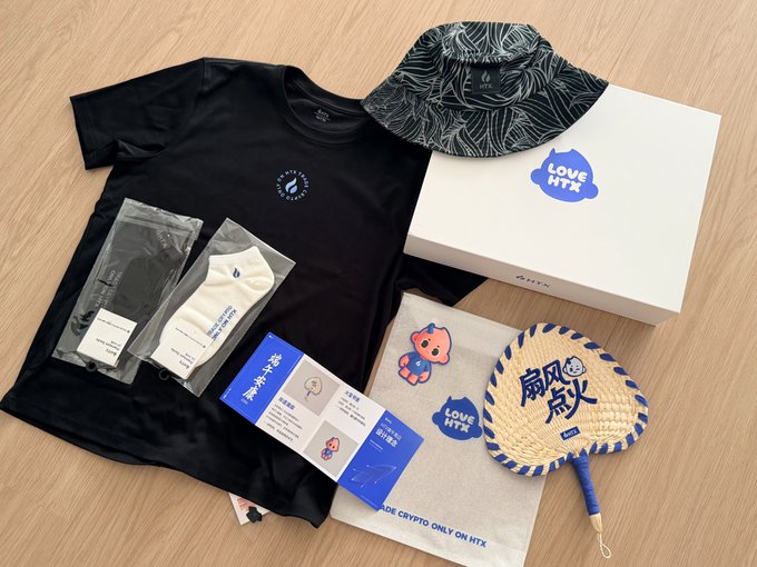 A flat-lay photo of a HTX (Huobi Global) Dragon Boat Festival gift box, featuring a black T-shirt, patterned bucket hat, socks, a cute Huobao bear sticker, and a traditional woven fan with "扇风点火" (fanning the flames) text alongside "LOVE HTX" branding.