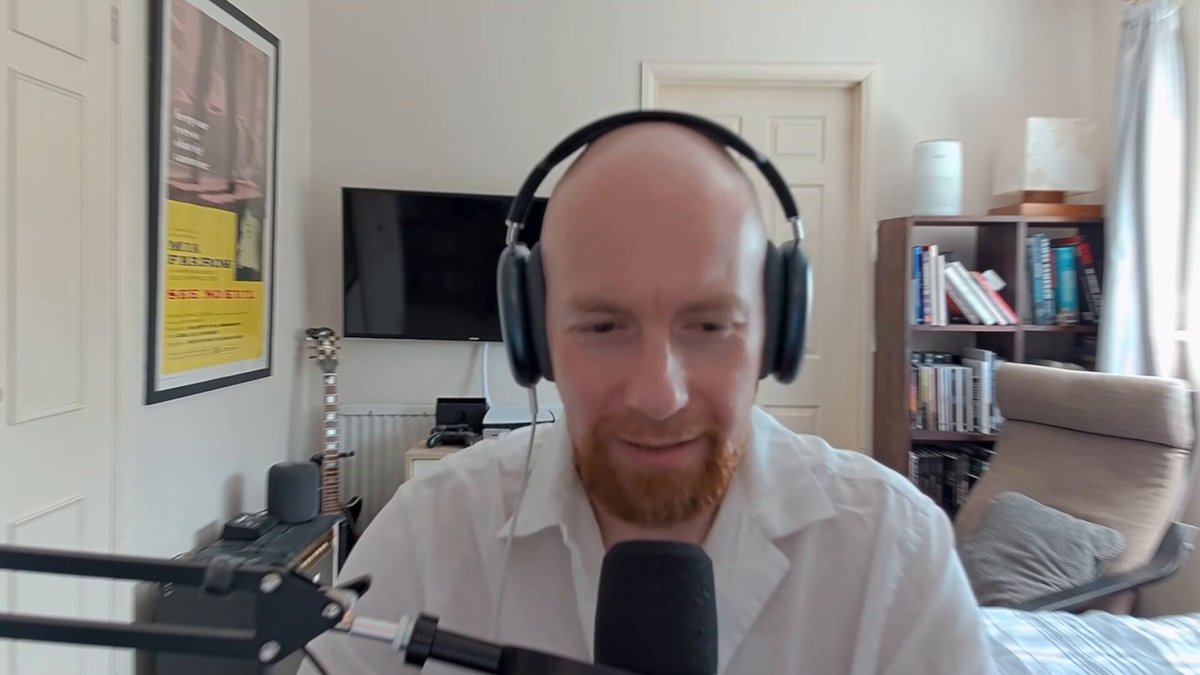 The image shows a man wearing headphones while sitting at a desk with a microphone. The man appears to be talking into the microphone or recording a video call using an app like Zoom. The background of the image features a TV screen, suggesting that he might be in a living room or a similar setting where multiple screens are present.