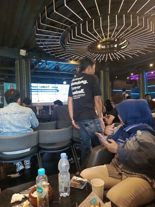 The image shows a group of people sitting around a table, with one person standing up holding a cell phone. The scene takes place at a restaurant or bar where there are chairs and dining tables visible. A water bottle can be seen on one of the tables, along with cups and other items like a book and a remote control.

The post is about a band called Bandung DeFi & Web3 that has been active for some time now. The image captures a moment where people are enjoying their time together at this event.