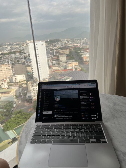 A silver laptop sitting on a desk by a window, displaying what appears to be Twitter/X on the screen, with a view of a city and mountains in the background.