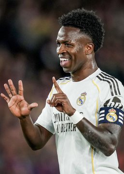 The image shows soccer player Vinicius Jr. in a Real Madrid kit, smiling and gesturing with his hands raised. The jersey features "Emirates Fly Better" sponsorship text and the club's crest, visually tying into the news of his new contract extension with Real Madrid through 2032.