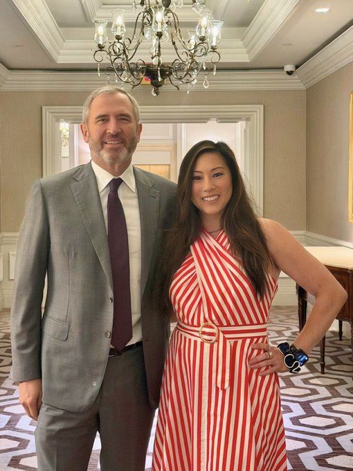 A man in a gray suit and purple tie stands beside a woman in a red and white striped halter dress with a gold D-ring belt, both smiling in an elegant room beneath a crystal chandelier. The image accompanies a post celebrating a long-term professional partnership in innovation, highlighting the progress they've made since 2022.