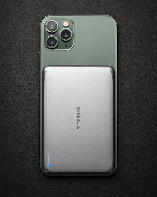 An iPhone with a silver TORRAS magnetic power bank attached to its back, displayed on a dark textured surface.