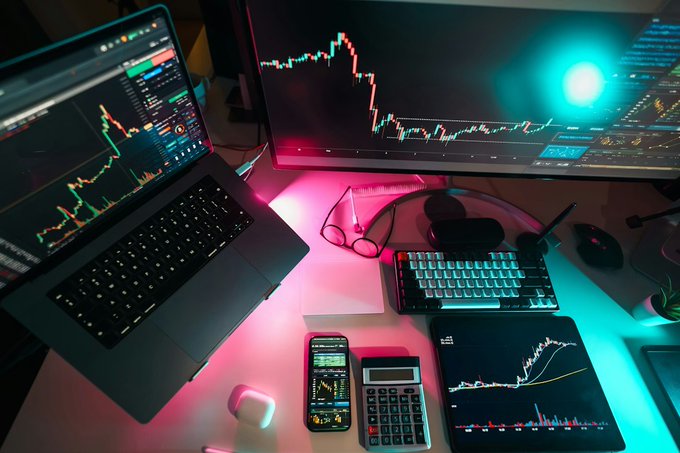 A dimly lit workspace features a laptop and an external monitor both displaying stock market candlestick charts, alongside a backlit mechanical keyboard, smartphone, calculator, eyeglasses, and a wireless mouse on the desk.