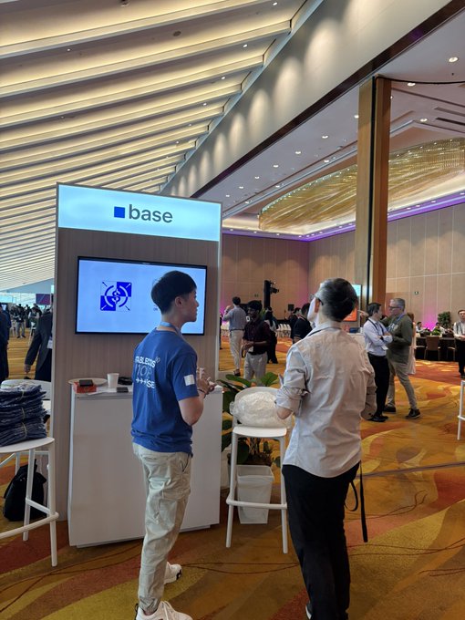 A booth displaying the "base" logo with a staff member in a blue "BUIDL INTO" shirt explaining the platform to an attendee at what appears to be a Singapore conference, illustrating the team's outreach to educate the TradFi space about Base in APAC.