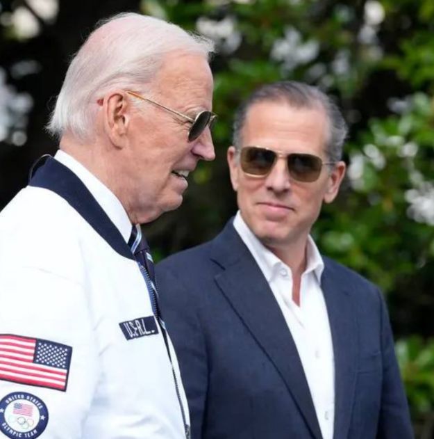 An older man wearing sunglasses and a white jacket with an American flag and 'U.S.A.' patch stands next to actor Hunter Biden, who is wearing a dark suit and sunglasses.