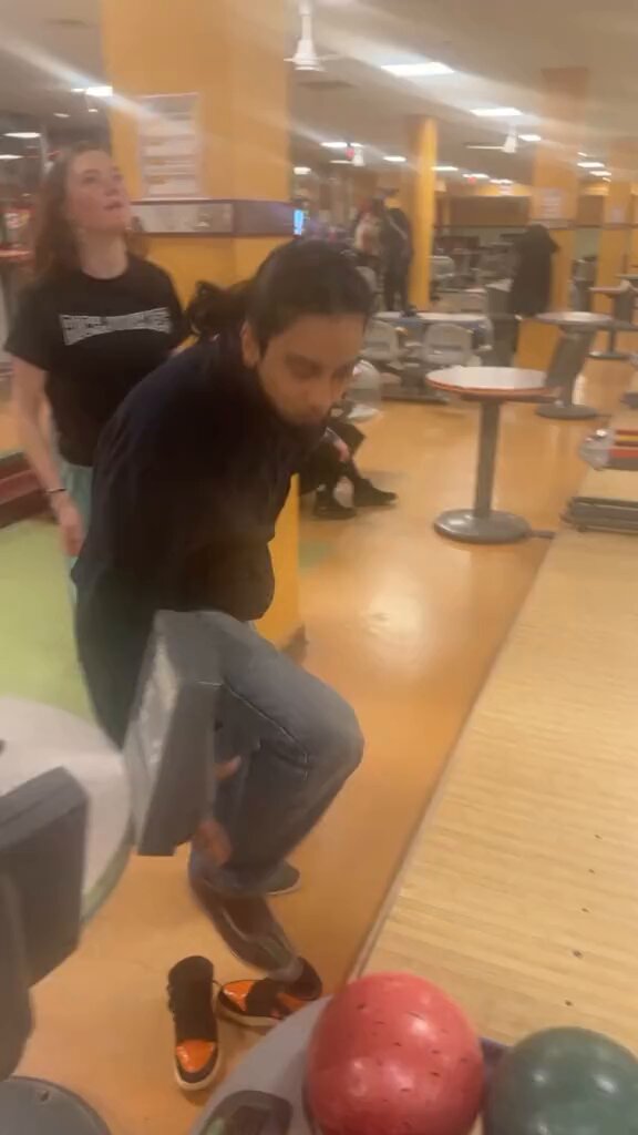 The image shows two people at a bowling alley, with one person bending over to pick up a red ball from the floor while another person watches nearby. There are also several other sports balls scattered around the scene, indicating that this might be a popular spot for various activities or games involving sports equipment. The post in the image reads "trying a long here", which suggests that someone is attempting to do a long jump or possibly a similar athletic feat at the bowling alley.
