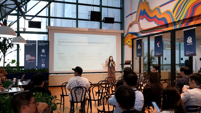 A woman presents to an audience in a modern room with colorful wall art, standing beside a projection screen displaying 'Models keep getting smarter. The internet still won't let them ADO.' The seated attendees are visible in the foreground, with banners reading 'Web3 BOOSTING' and 'Opens Up About About Us' in the background.