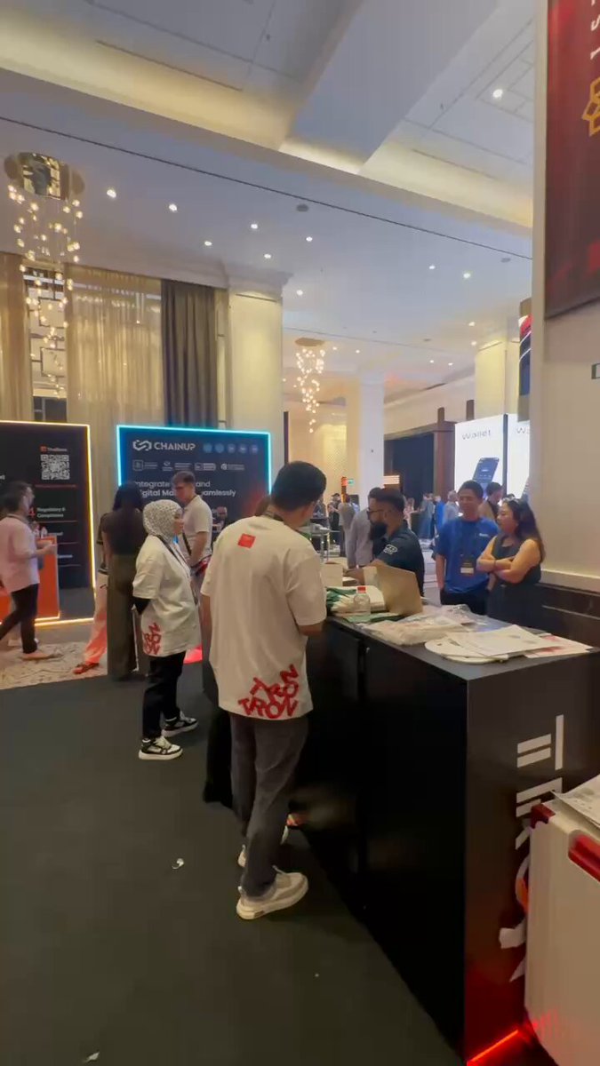 The image shows a group of people standing around a booth at an event. There are several individuals present, including one person wearing a white shirt with red designs. The booth appears to be selling something or providing information about a product or service. A man is standing behind the booth and talking to some attendees, while others look on from various positions within the scene.