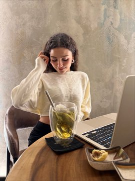 A young woman with curly hair sitting at a wooden table in a cafe working on a laptop, with a cup of tea and a pastry beside her.