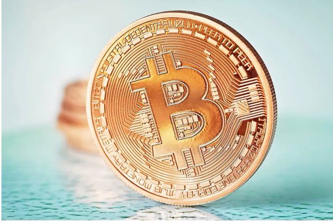 A close-up shows a gold-colored Bitcoin coin standing upright on a textured teal surface.