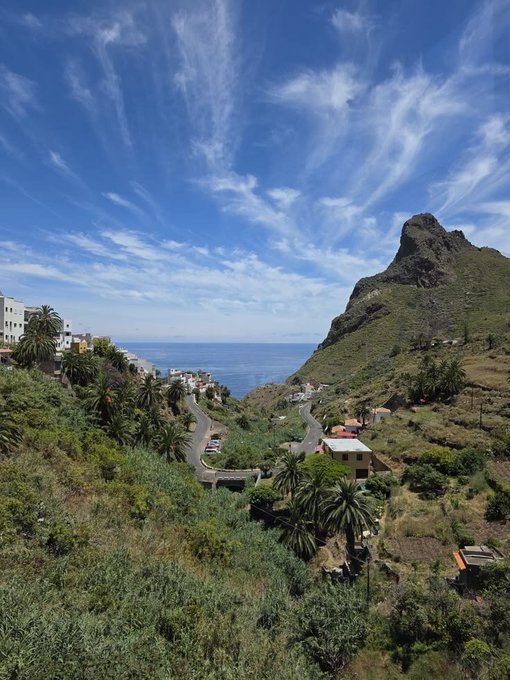 A scenic landscape view features a winding road descending through a lush, green valley filled with palm trees and white houses, leading toward the ocean, with a prominent rocky peak on the right side under a partly cloudy blue sky.