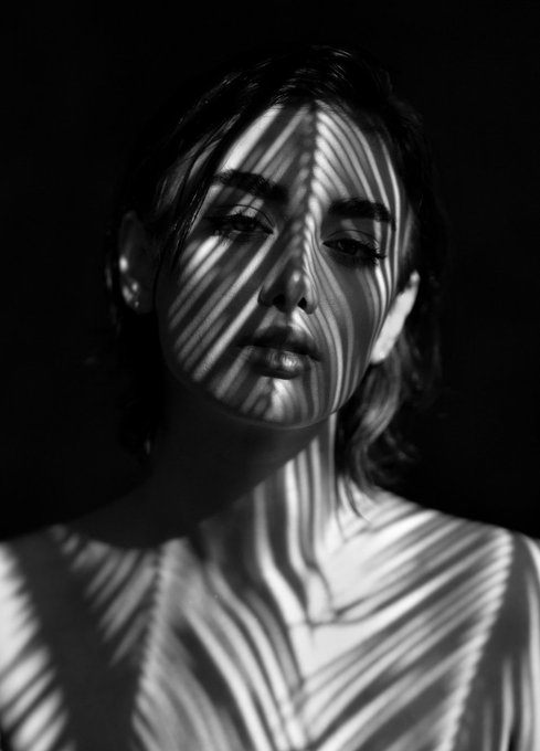 A high-contrast black-and-white portrait photograph of a young woman's face and neck, illuminated by striking, wavy linear light patterns against a dark background.