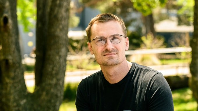 A man with glasses and short hair, wearing a black t-shirt, sits outdoors in a sunlit park setting, looking directly at the camera with a serious, slightly stern expression. His somber demeanor visually embodies the post's sentiment about "Rune" being upset, conveying a quiet frustration or displeasure through his downturned mouth and intense gaze.