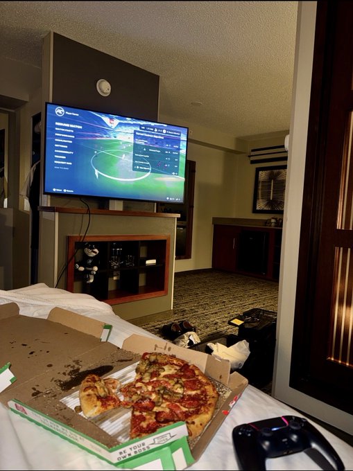 A hotel room scene showing a TV displaying what appears to be a soccer broadcast menu, with an open Papa John's pizza box on the bed in the foreground.