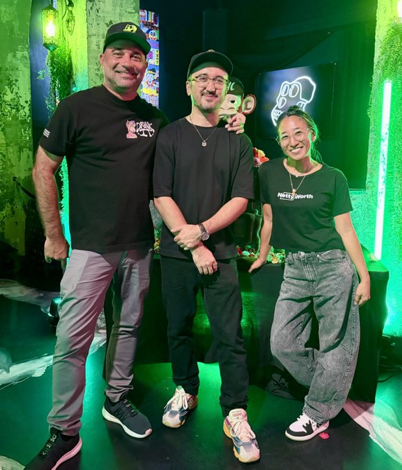 Three people posing for a photo in a room with green lighting; one man wears a "SMASH NYC" shirt, and the woman wears a "Nifty Worth" shirt.
