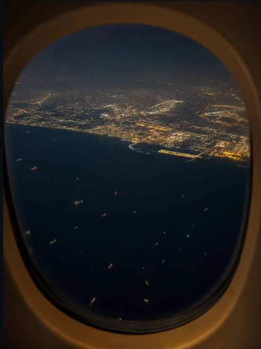 A nighttime aerial view of a brightly lit coastal city and harbor is seen through an airplane window oval, with ships dotting the dark waters below.