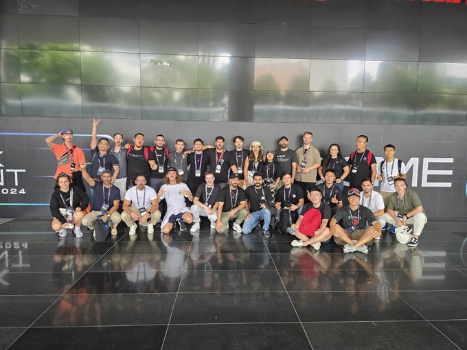 A large group of conference attendees poses for a team photo in front of a dark wall with partial "TIME" lettering, with one person in the back giving a peace sign.