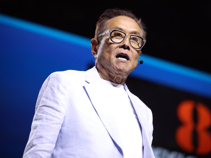 The image features a man wearing glasses and a white suit standing on stage with his mouth open as he speaks into a microphone. The background of the image shows a blue wall, which could be part of an event or presentation. There are also some words visible in the image that seem to be related to the post by Robert Kiyosaki.