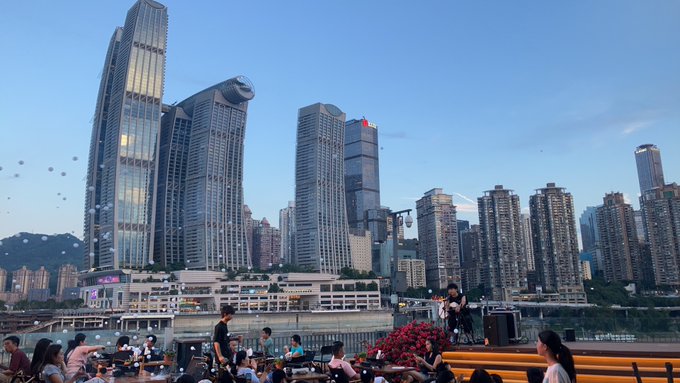 A photograph of a dense city skyline at dusk featuring modern skyscrapers, including one with 'PLAZA' signage, with people gathered at a viewpoint in the foreground.