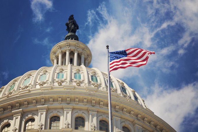 A low-angle shot of the U.S. Capitol dome against a blue sky with clouds, with an American flag waving on a flagpole in the foreground.