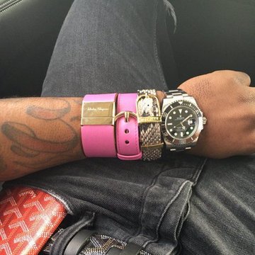 A close-up of a tattooed wrist displaying three watches: a pink-banded watch with gold accents, a jeweled watch, and a silver Rolex Submariner, resting on dark denim.