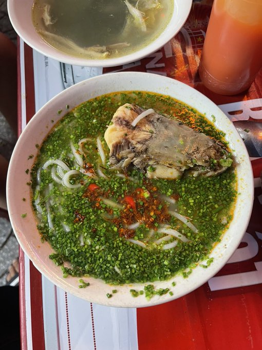 An overhead view of a bowl of noodle soup topped with chopped green herbs, red chili flakes, and a large piece of meat (possibly bone-in chicken or fish), accompanied by a smaller bowl of clear broth and a glass of orange-colored drink.