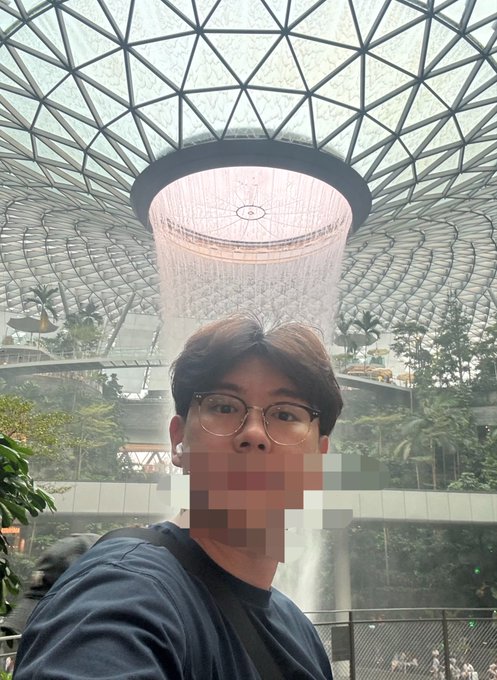 A selfie of a young man wearing glasses at what appears to be Jewel Changi Airport in Singapore, with the iconic Rain Vortex waterfall visible behind him.