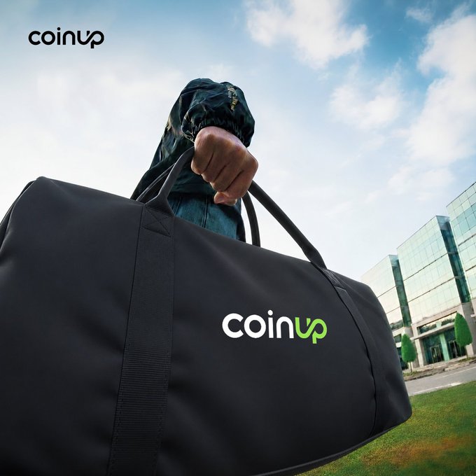 The image shows a person holding a black duffle bag with "coinup" written on it. The person appears to be walking or standing outside, possibly at an office building. The scene suggests that the individual might be preparing for work or heading out for a day of activities involving their coinup account.