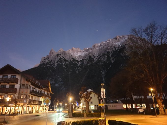 A twilight view of a snow-capped mountain towering over a quaint Alpine village, with warm streetlights illuminating traditional chalets and a quiet town square. The "GN fam" caption likely refers to "Garmisch-Nacht" or a group visit to Garmisch-Partenkirchen, the famous Bavarian ski town known for its dramatic mountain scenery like the Zugspitze massif shown here.