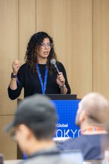 A woman with curly hair speaks into a microphone at a podium displaying a 'Global' and 'QKY' logo.
