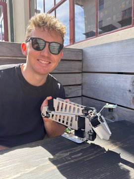 A smiling young man with curly blonde hair and sunglasses sitting outdoors on a wooden deck, holding a white robotic arm device.