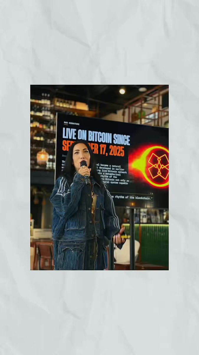 The image features a woman standing next to a poster or sign with the words "Live on Bitcoin" written on it. The poster also includes an orange flower design that adds visual interest. The background of the image shows a dining table, suggesting that this event might be taking place in a restaurant or similar setting.