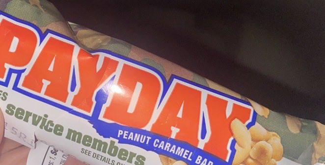 A close-up photograph of a PayDay peanut caramel bar wrapper, showing the text "PAYDAY," "PEANUT CARAMEL BAR," and "service members."