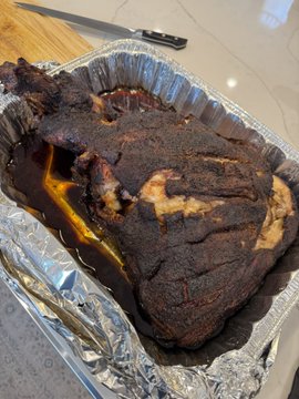 A large, well-burnt brisket rests in a foil pan with rich juices pooling around it, alongside a knife, showcasing the smoky results of a successful barbecue cook.