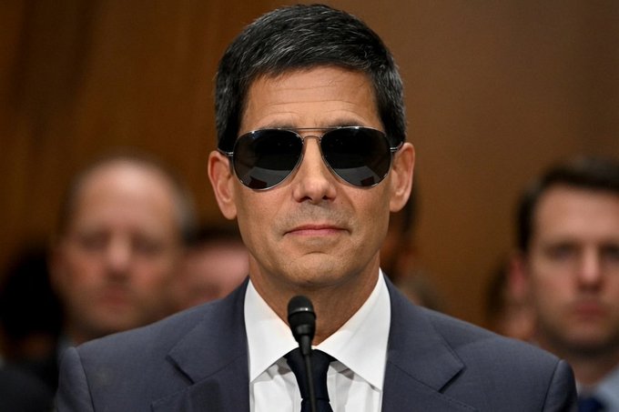 A man in a dark suit and sunglasses sits at a microphone in what appears to be a formal hearing or conference setting, with blurred figures visible in the background. The image's serious, official atmosphere contrasts humorously with the post's optimistic caption about a bullish cryptocurrency chart for XRP/Ripple, suggesting a meme-like juxtaposition of financial optimism with a stern demeanor.