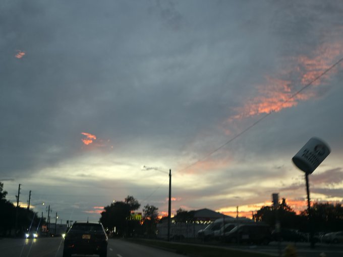 A dusky sunset view from a car dashboard, showing a sky with pink-tinged clouds over a road with vehicles, power lines, and a roadside sign.