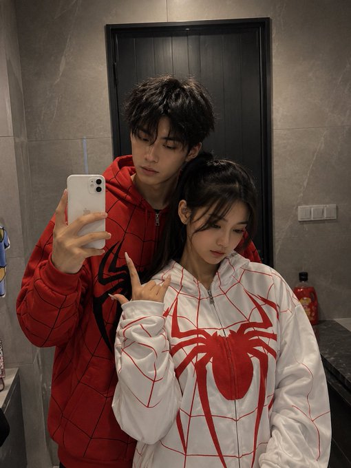 A couple poses for a mirror selfie wearing matching Spider-Man-themed hoodies, with the man in red and the woman in white featuring a red spider emblem.